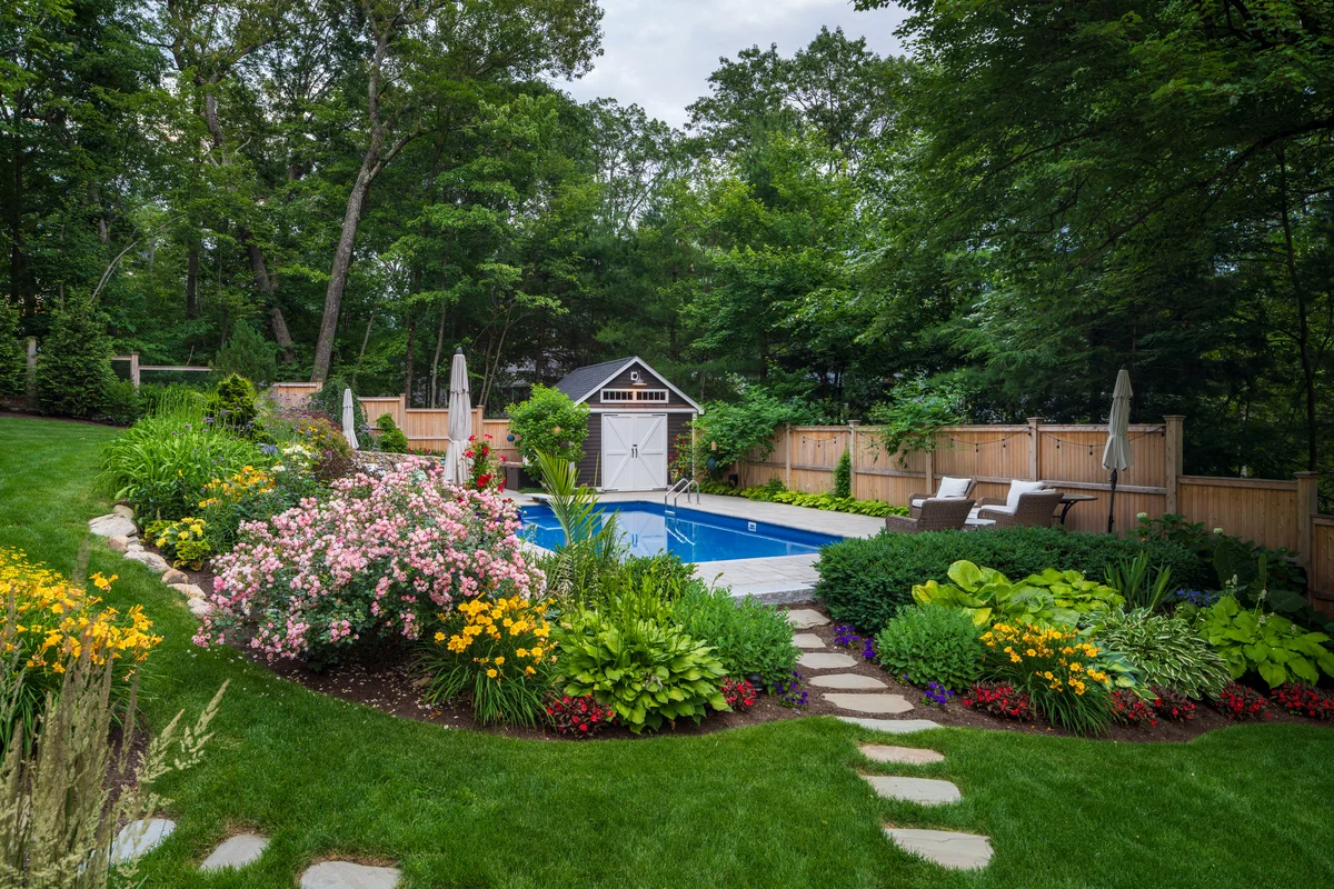 Custom backyard pool and landscape design in Middlesex County MA with vibrant flower beds, stonework, and patio seating.